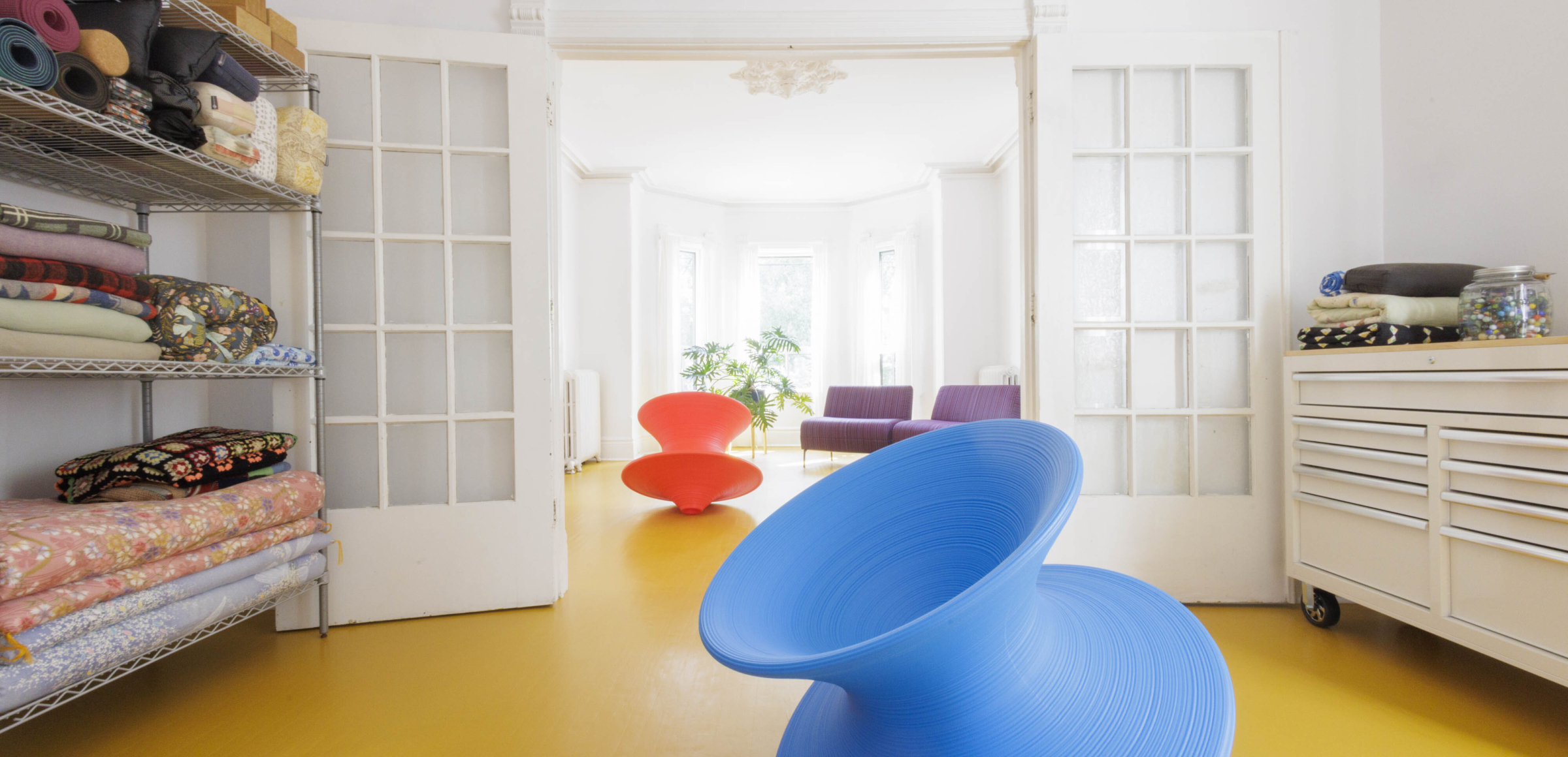 The Sensory Studio: view through the two expansive adjoining rooms, french doors, shelf with sensory props, red and blue spun chairs, philodendron and welcoming purple couch by light filled bay windows.