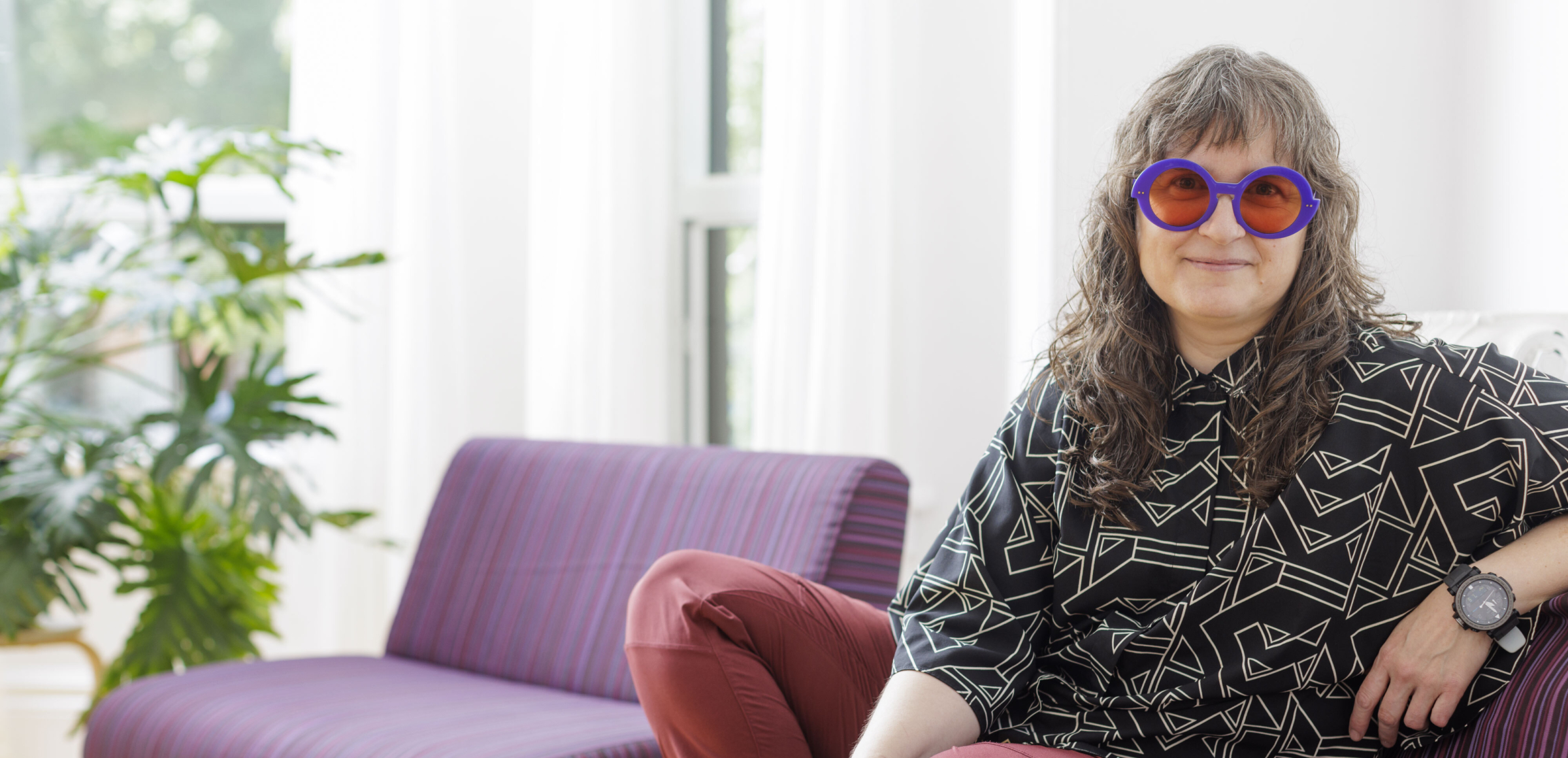 Ayasha sitting cross legged on the purple couch with funky new wave shirt and rose tinted glasses.