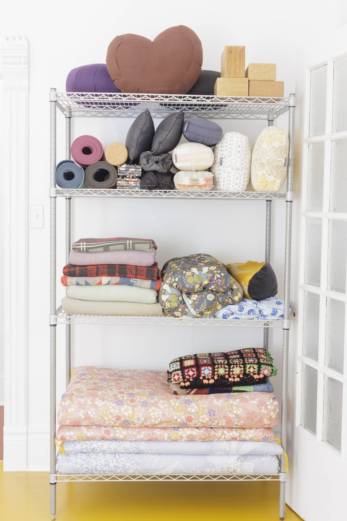 Sensory, movement and somatic props arranged playfully on a light grey wire shelf. Big brown heart pillow, bolsters, mats, cork blocks, blankets and shikibutons.